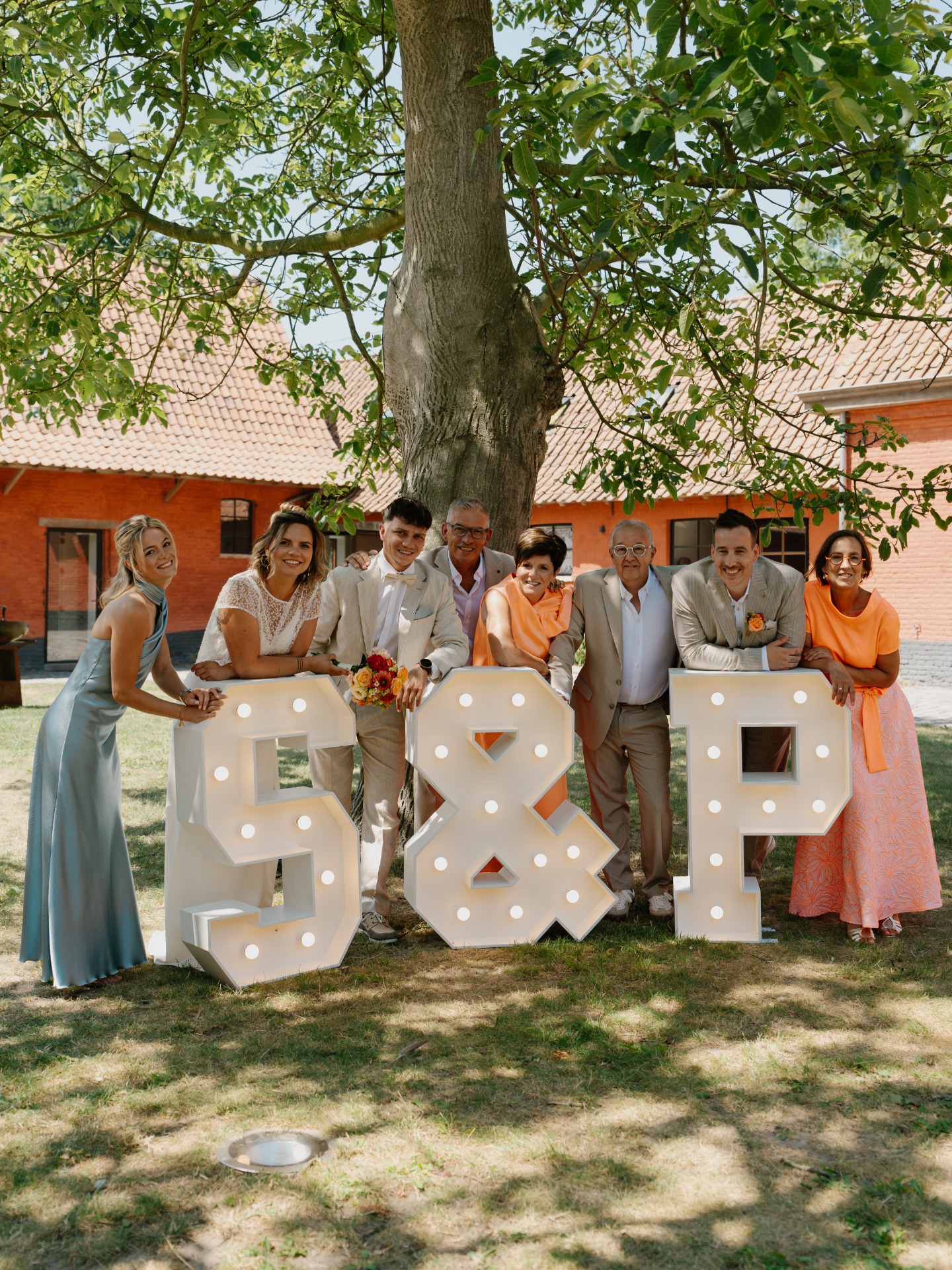 Groep mensen poseert buiten onder een grote boom, met verlichte witte letters “LOVE” op het gras.